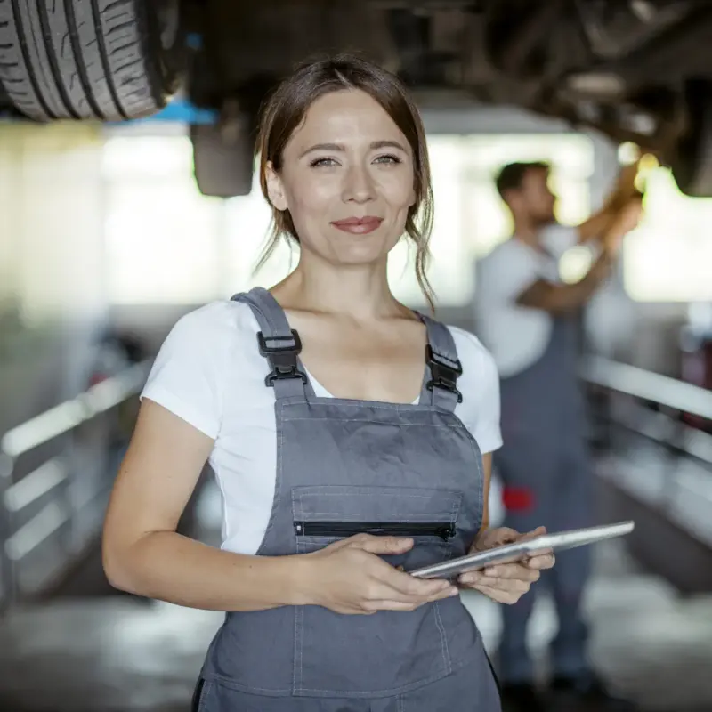 A smiling female mechanic in gray coveralls holds a tablet in a repair shop while a coworker works beneath a car raised on a lift behind her.