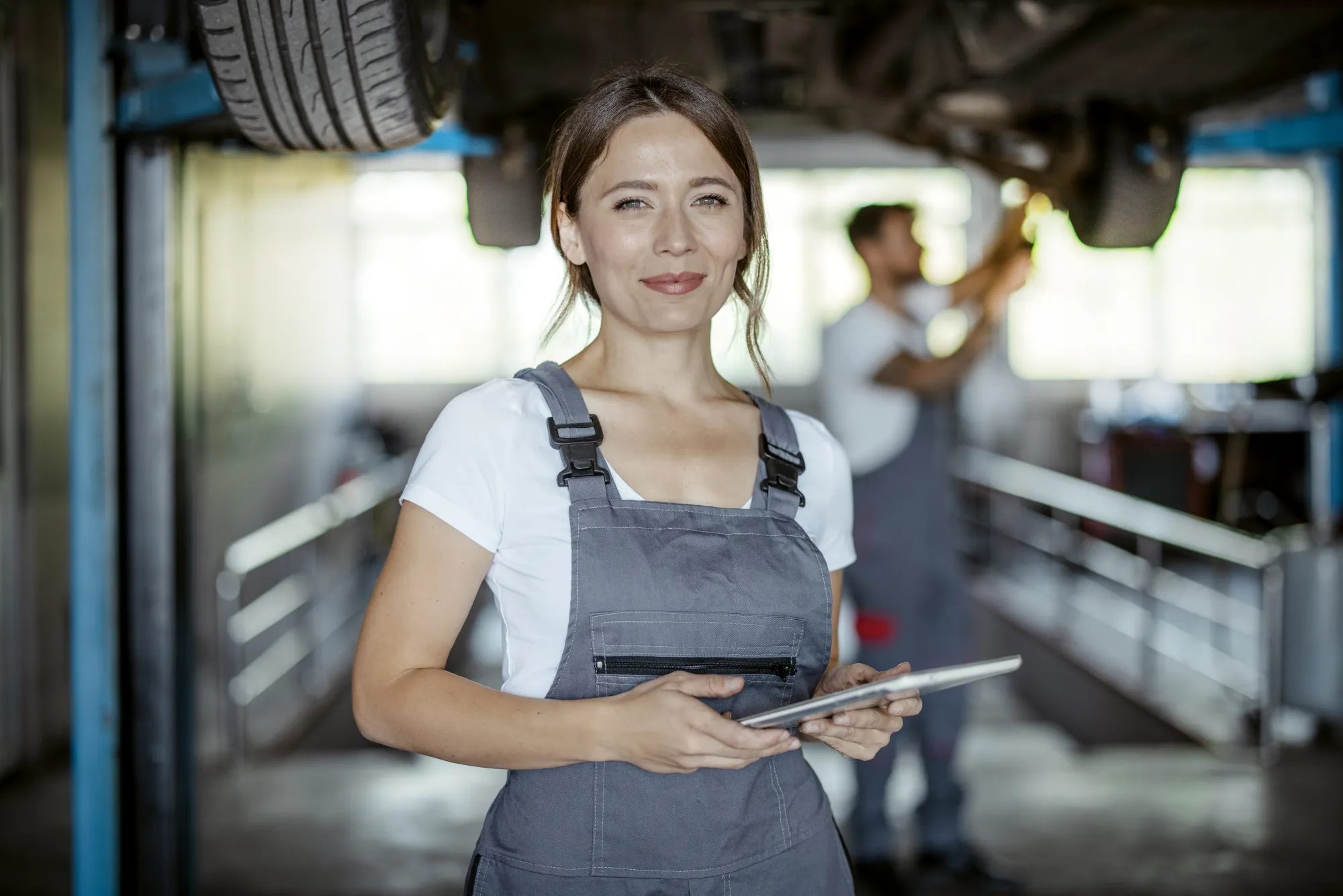 A smiling female mechanic in gray coveralls holds a tablet in a repair shop while a coworker works beneath a car raised on a lift behind her.