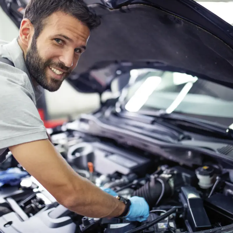 A smiling bearded mechanic in a grey shirt reaches into the engine bay of a car with its hood propped open inside a repair shop.