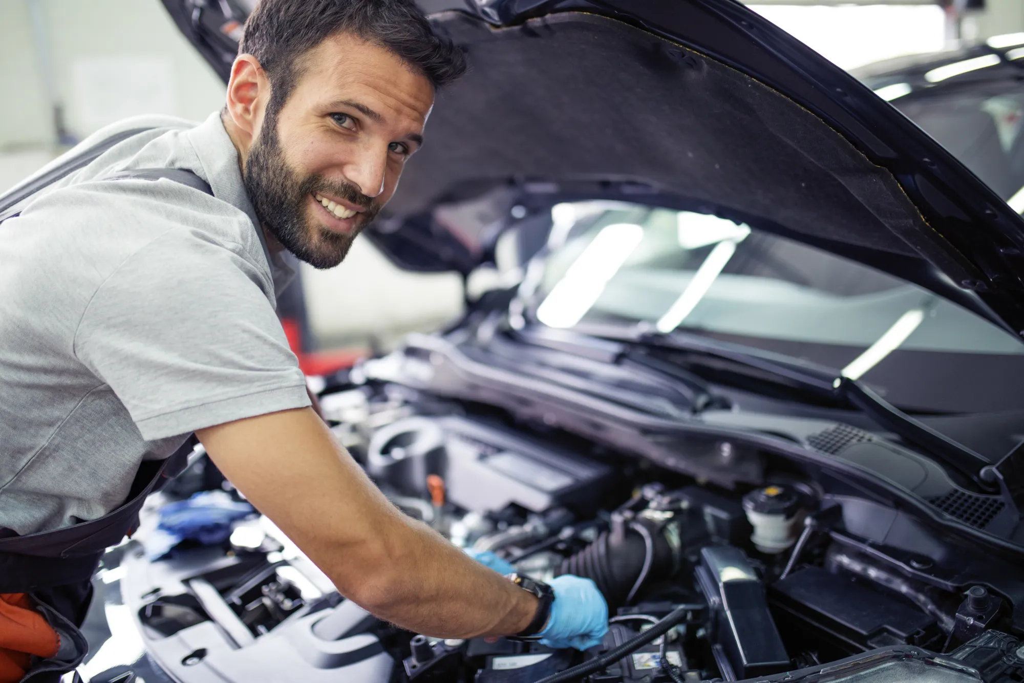 A smiling bearded mechanic in a grey shirt reaches into the engine bay of a car with its hood propped open inside a repair shop.